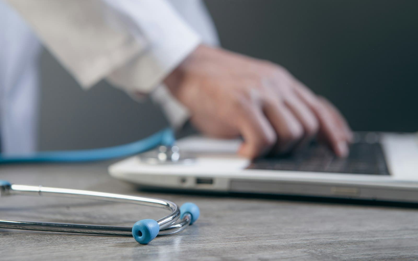Doctor in a white coat typing on a laptop, with a stethoscope lying on the desk in the foreground